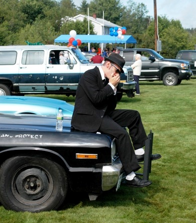 Blues Brother playing harmonica on car hood.